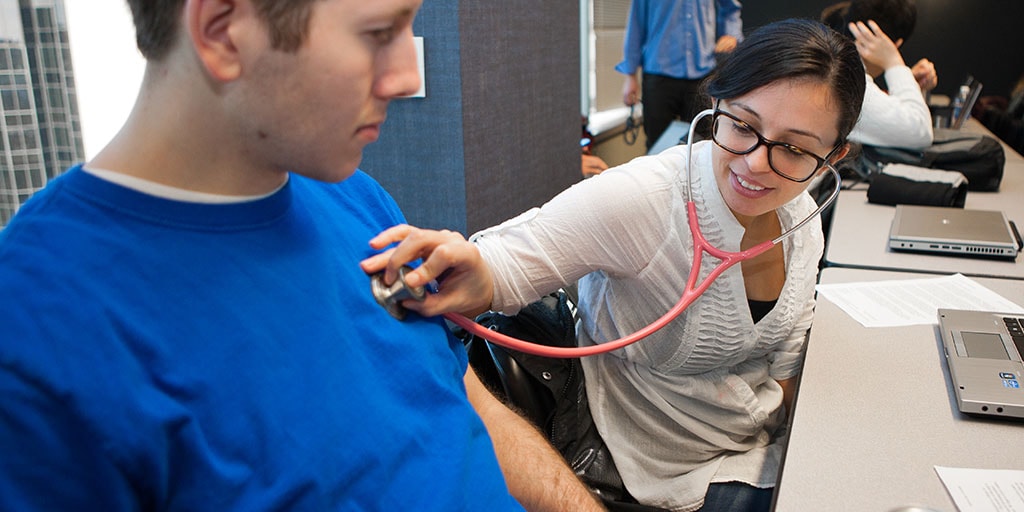 Two students in a classroom, one student is listening to the other student's heart with a stethoscope.
