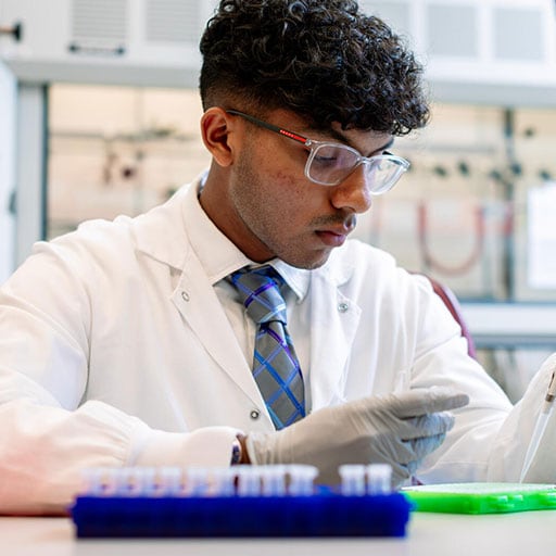 Young research student in a lab coat sitting in a lab setting, pipetting something into a dish