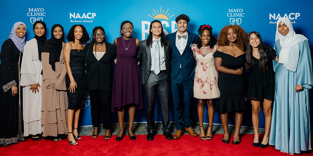 RISE for Youth students and mentors gather together for a group photo in front of a blue wall with Mayo Clinic and NAACP logos.