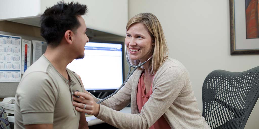 An NP smiling at a patient and checking their heart beat in an exam room setting