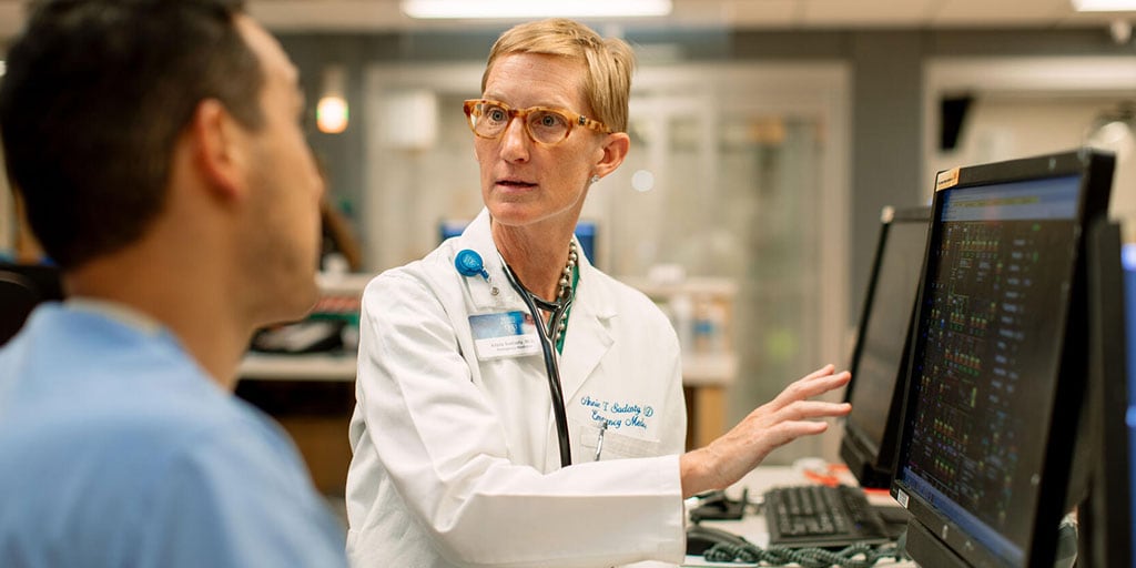 A faculty member wearing a white coat pointing to something on a computer screen while talking to a fellow.