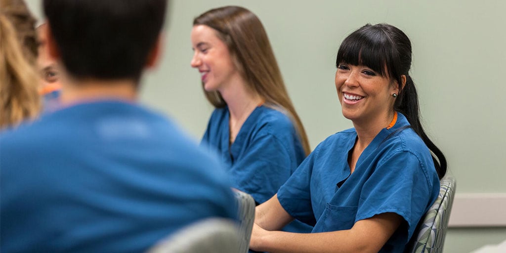 Three students sitting at a table in a classroom. Two are looking towards the front of the room, one is looking towards the camera, smiling.