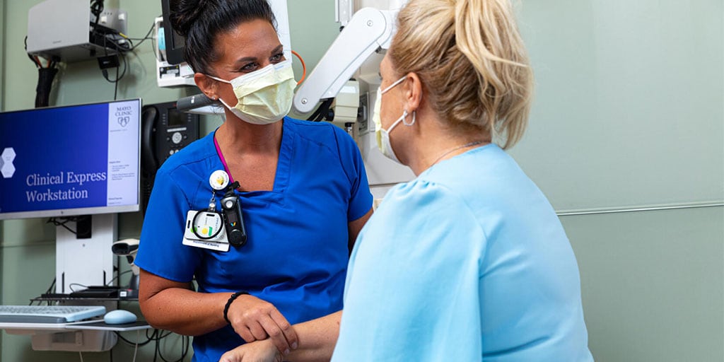 A nurse and patient standing in an exam room, facing each other. The nurse is taking the patient's pulse.