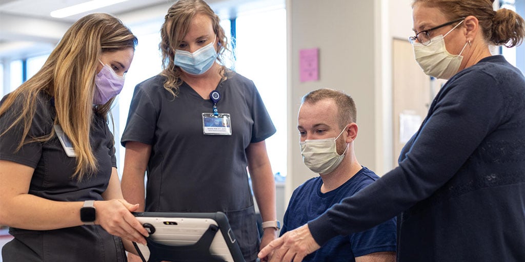 A group of 4 people wearing masks and blue scrubs, grouped around and reviewing an ipad