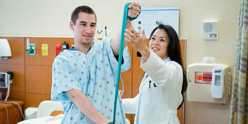 OT Provider and patient in a gown, both in a patient room doing hand strength exercises.