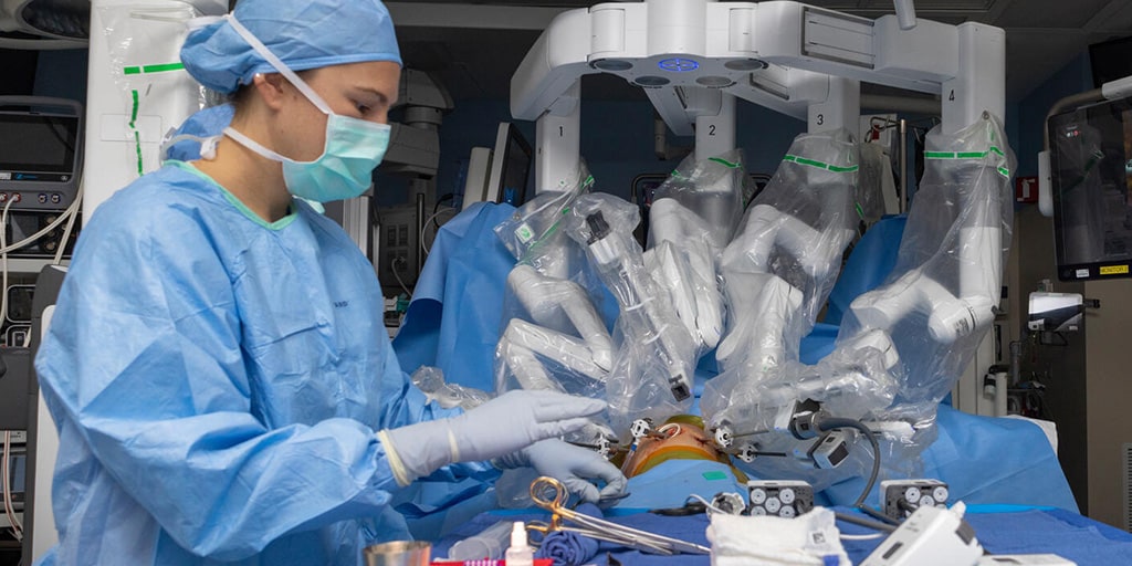 A member of a surgical care team attends the site of a surgical procedure being performed with a robotic surgical system, controlled remotely by a surgeon at a console.