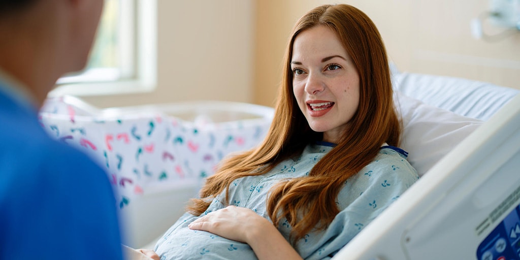 A pregnant woman in a hospital bed talks with a nurse.