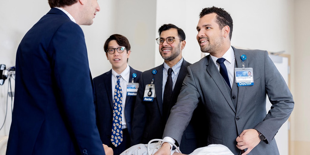 A nephrology fellow practices performing an ultrasound on a medical mannequin while his colleagues are gathered around and view the screen.