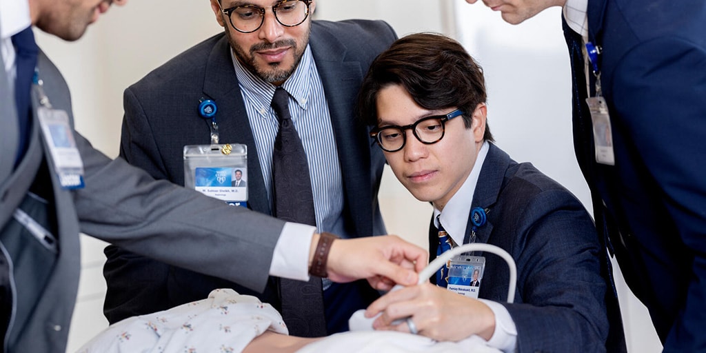 A nephrology fellow practices performing an ultrasound on a medical mannequin while his colleagues are gathered around.