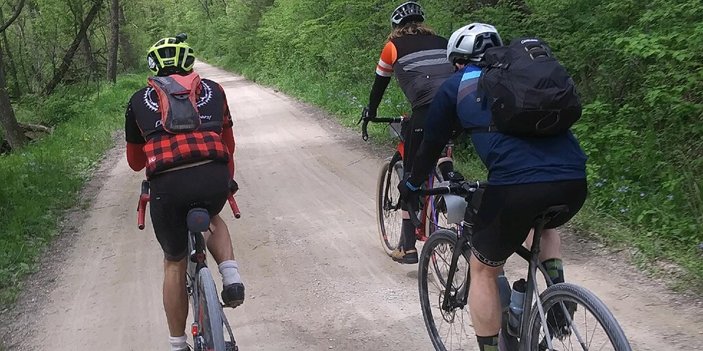 A group of Mayo Clinic residents rides bicycles along a wide, packed dirt trail bordered by lush green trees and dense foliage. All three cyclists wear helmets and backpacks, and they are shown from behind as they travel together on a quiet, wooded path near Rochester, Minnesota.