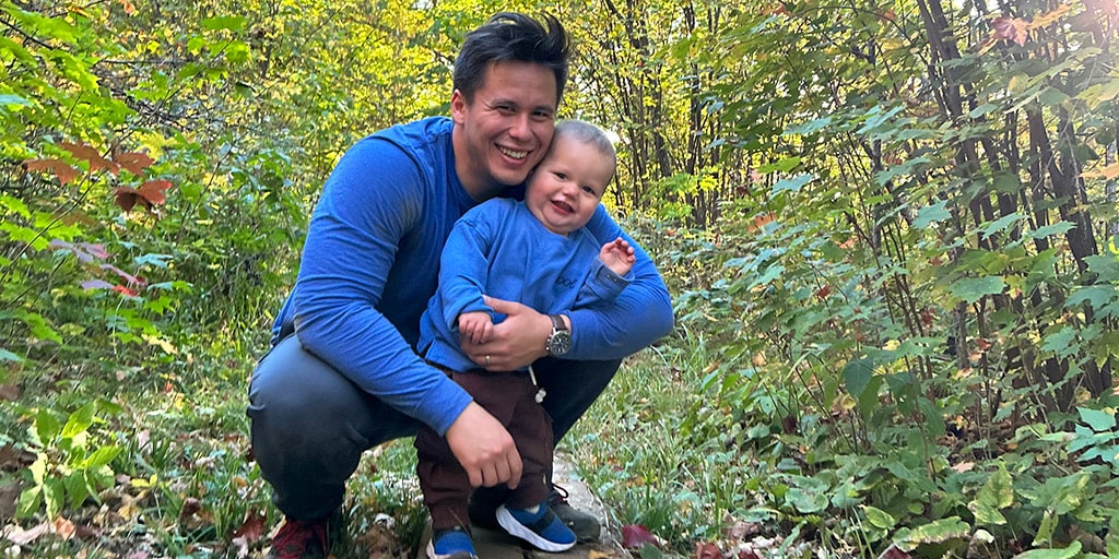 Neurosurgery resident Jeremiah Harrison crouches on a wooden boardwalk in a forest at Quarry Hill, smiling as he wraps an arm around his young son. They’re both dressed in blue, surrounded by green foliage and scattered autumn leaves, enjoying a quiet moment together on the trail.