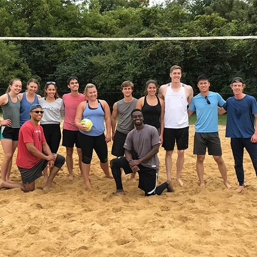 Group of students on a sand volleyball court, all looking at camera and smiling